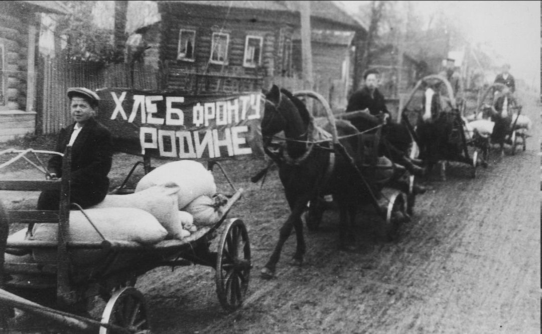 ХЛЕБ ФРОНТУ РОДИНЕ. На фотографии изображена колонна конных повозок, груженных мешками, вероятно, с зерном или другими продуктами. Люди управляют повозками. На переднем плане на одной из повозок сидит молодой человек. На заднем плане виден транспарант с надписью.