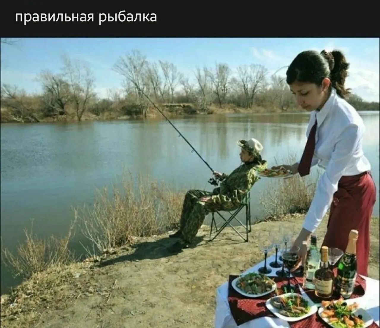 правильная рыбалка. Мужчина в камуфляже сидит с удочкой у реки, а рядом официантка накрывает стол с едой и напитками.