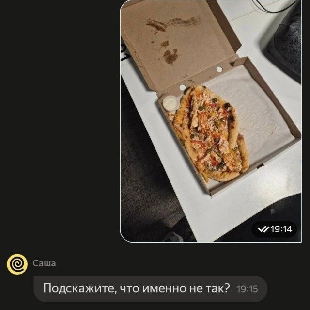 На изображении показана открытая коробка из-под пиццы, внутри которой лежит сильно деформированная, скомканная пицца, занимающая лишь часть пространства. Рядом с пиццей находится небольшой контейнер с соусом. На крышке коробки видны жирные пятна. Ниже изображения в интерфейсе чата (предположительно, Telegram) от пользователя «Саша» написано