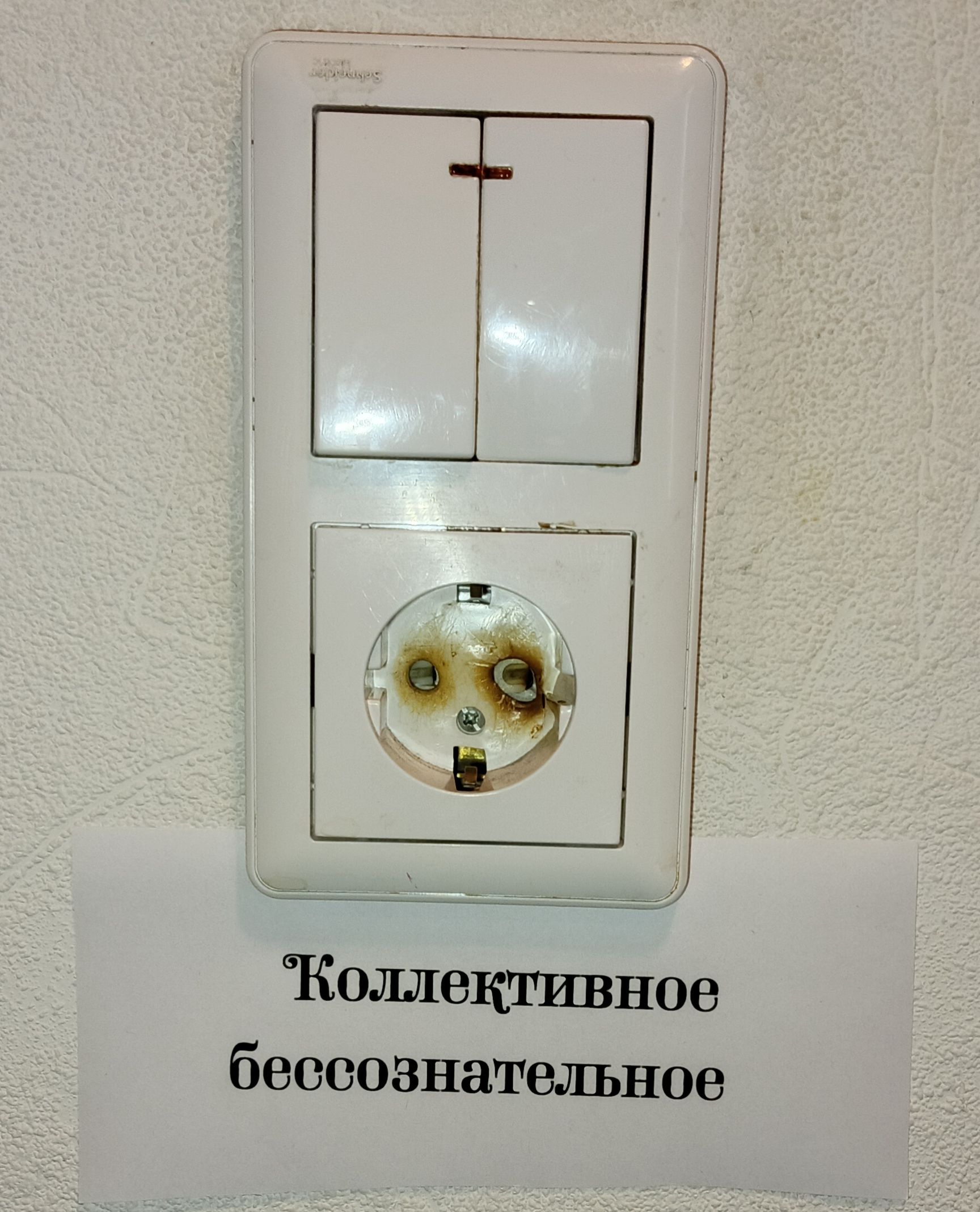 На стене висит белая розетка и выключатель. Розетка сильно обгоревшая и поврежденная. Под ней на листе бумаги написано: Коллективное бессознательное.