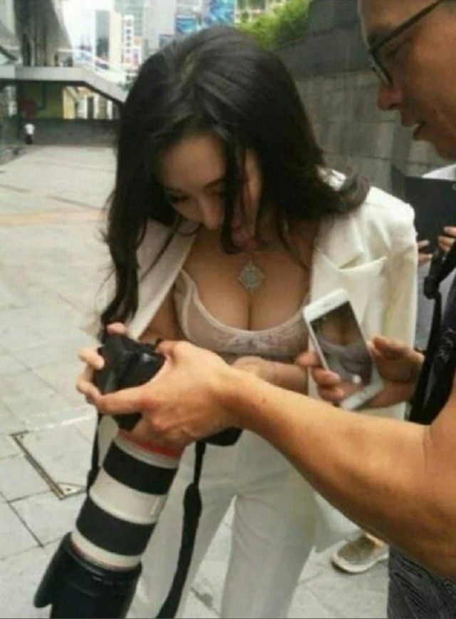 Женщина в белом пиджаке и прозрачном топе смотрит на камеру, которую держит мужчина. Другой мужчина держит телефон, на котором видно крупный план ее декольте.