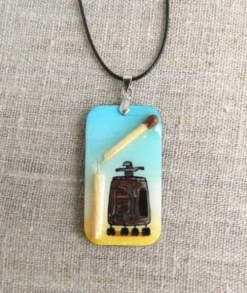 На подвеске изображён стилизованный рисунок на фоне градиента: сверху голубое небо, снизу жёлтая земля. В центре — неясный механизм или прибор, изображён в темном цвете.