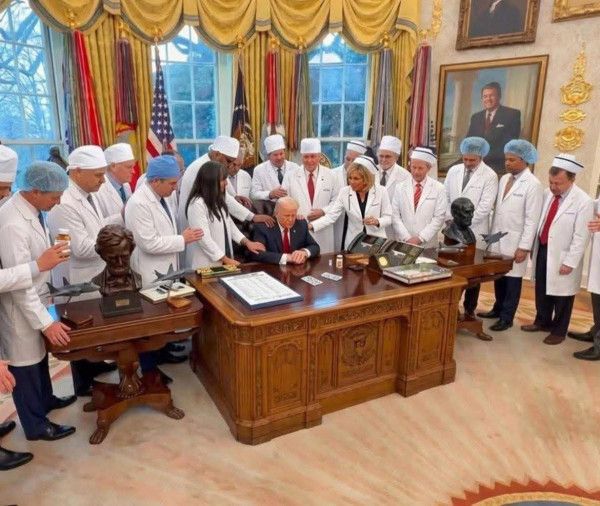 Групповой снимок вокруг большого письменного стола: люди в белых халатах и головных уборах стоят вокруг, главный подписывает документ в центре, в роскошной обстановке с портретами и занавесками.