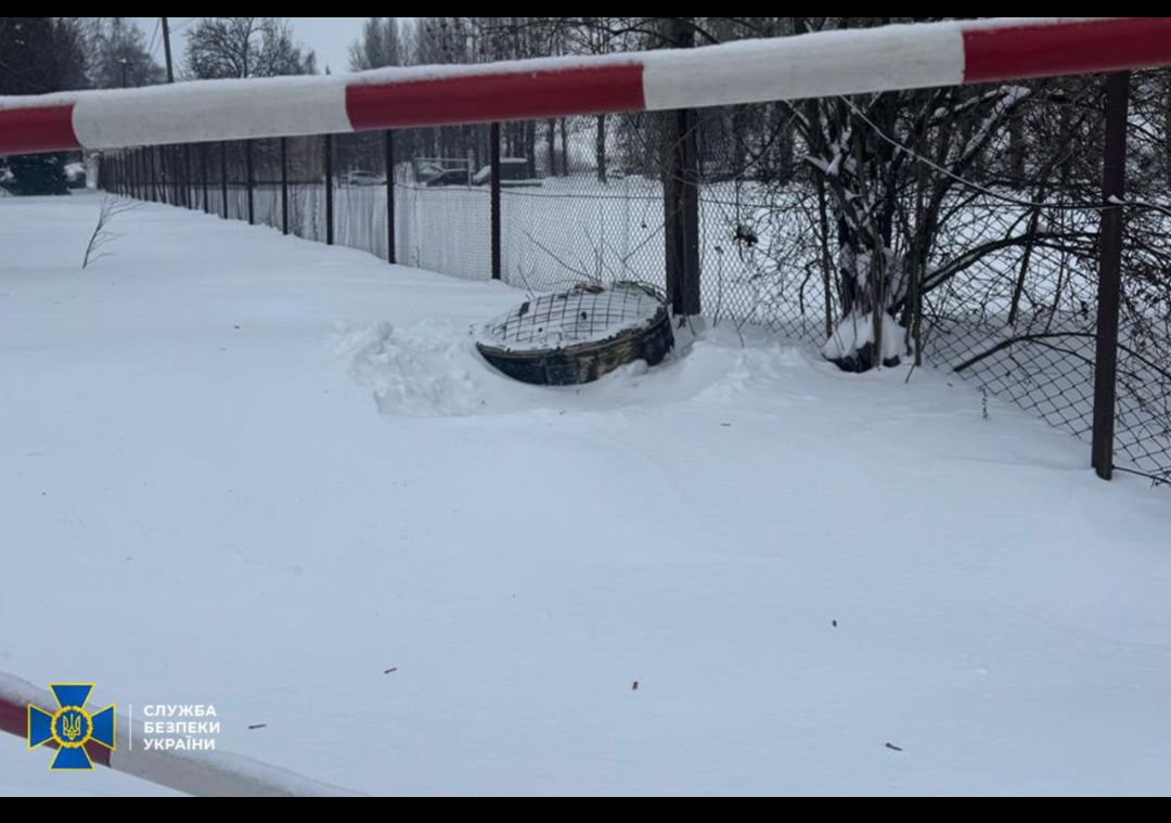 Заснеженная территория возле забора, рядом лежит закрытая крышка люка под охранной лентой.