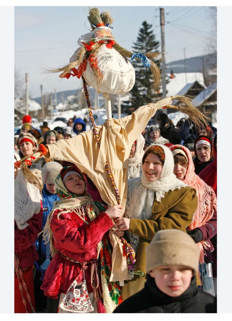Группа людей в народных костюмах на снежном празднике держит соломенное чучело.