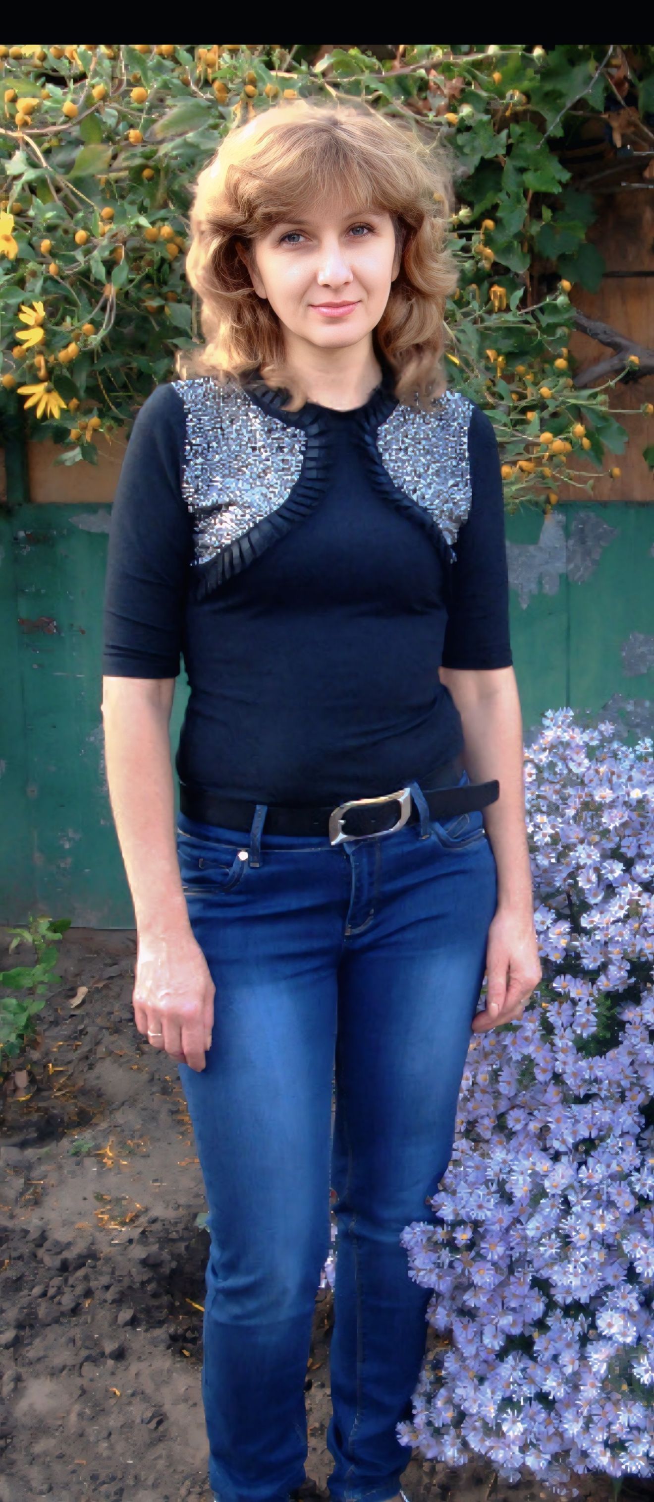 Женщина в тёмной кофте с блестящими вставками и джинсами стоит на улице возле кустов