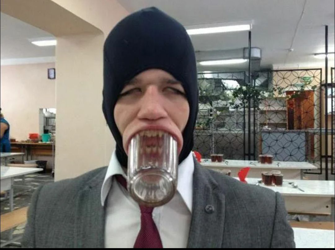Человек в костюме с балаклавой держит стакан прямо между зубами, создавая странный грим.