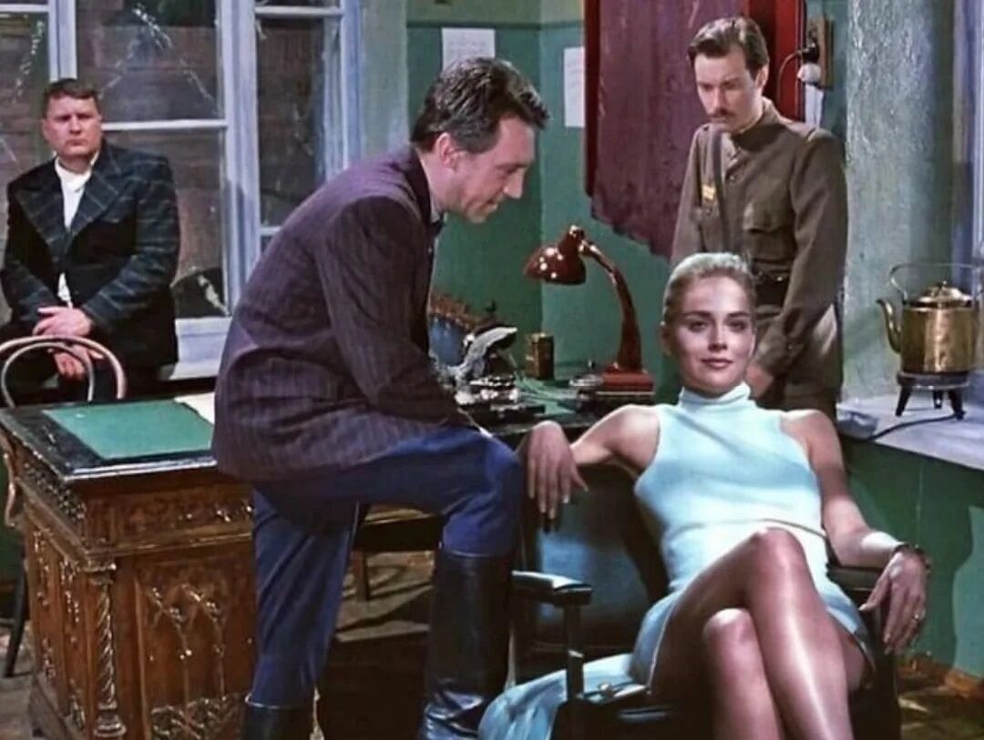 Сцена в ретро-офисе: мужчина беседует с женщиной в светлом платье, она сидит раскованные ноги.