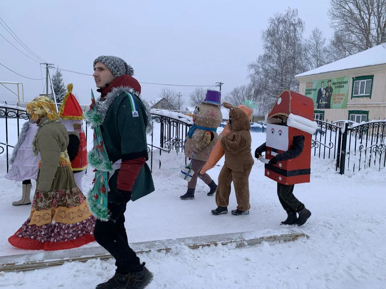 Группа людей в ярких костюмах идёт по снежной дороге на фоне зимнего двора.