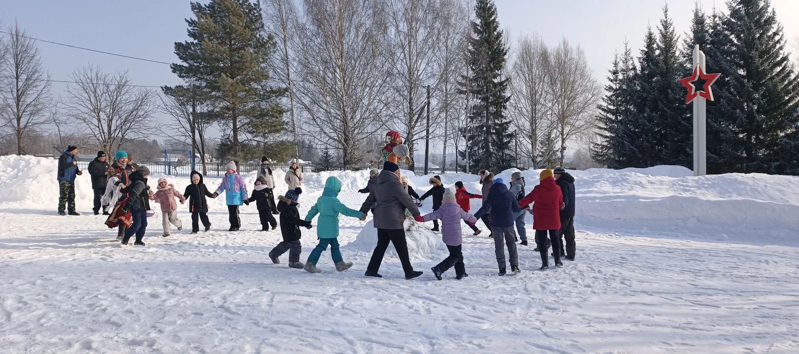 Группа людей держится за руки и образует круг на заснеженном поле; на фоне деревья и памятник с красной звездой.