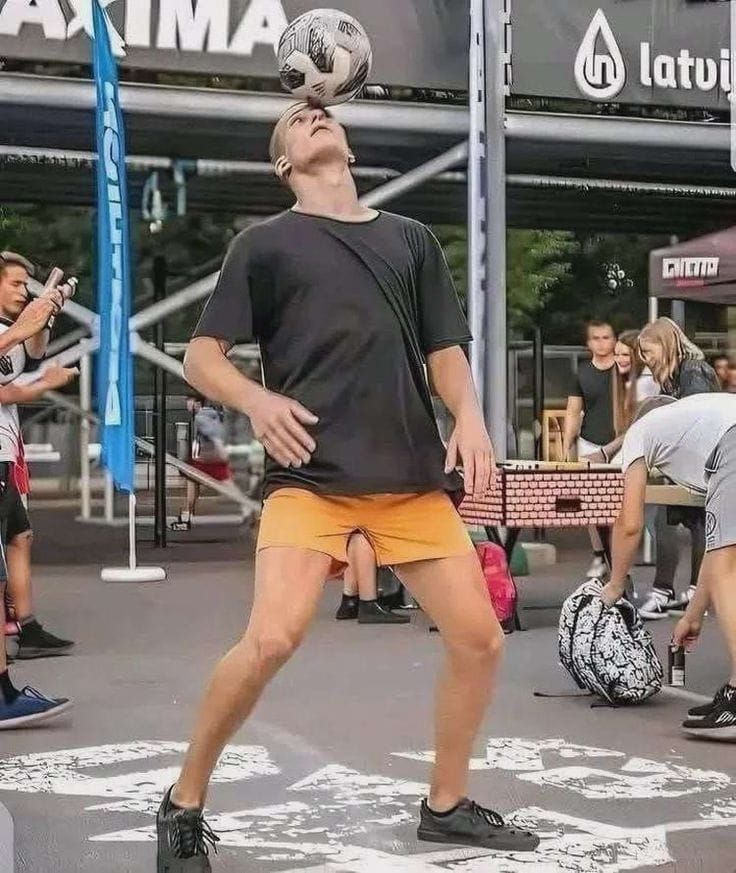 Мужчина балансирует мячом на лбу во время уличного мероприятия, вокруг собираются люди.