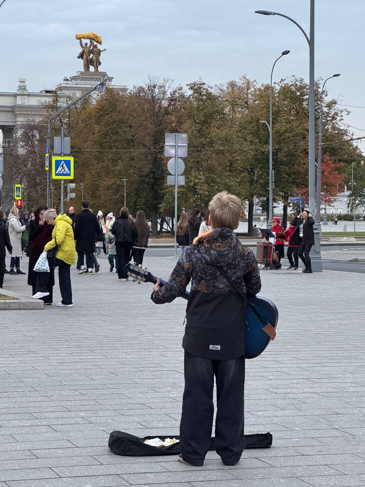 Уличный музыкант с гитарой на площади, за ним толпа прохожих и памятник вдалеке.