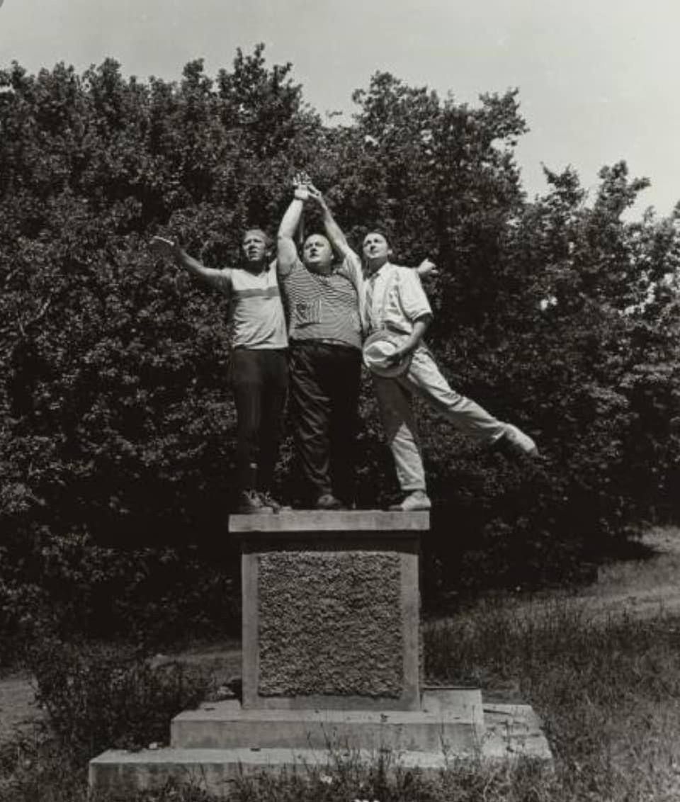 Трое мужчин стоят на монументе, держатся за руки и поднимают их вверх, один мужчина держится на одной ноге.