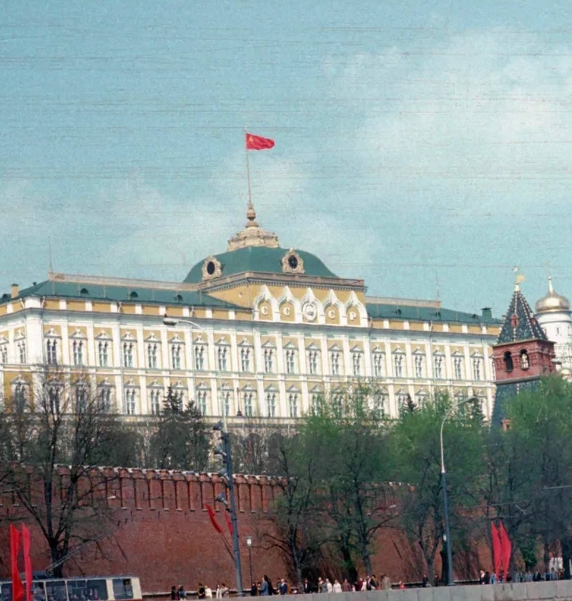 Большой административный дворец с красным флагом на крыше, вокруг деревья.