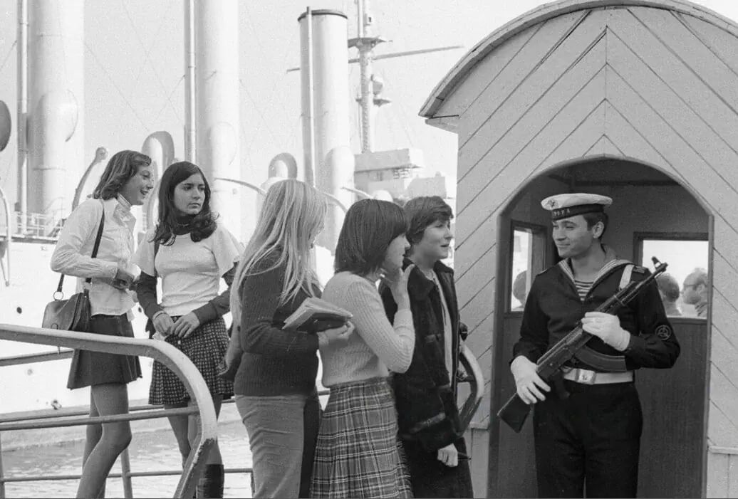 На пирсе группа девушек разговаривает с военнослужащим на корабле.