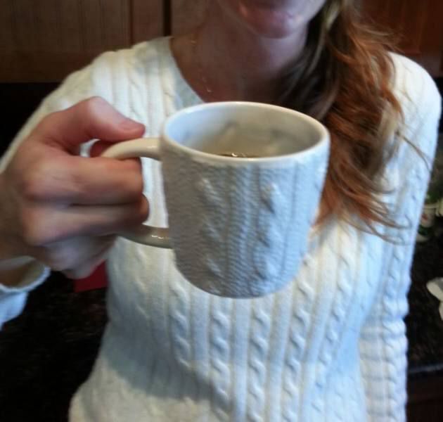 Человек в светлом вязаном свитере держит кружку.