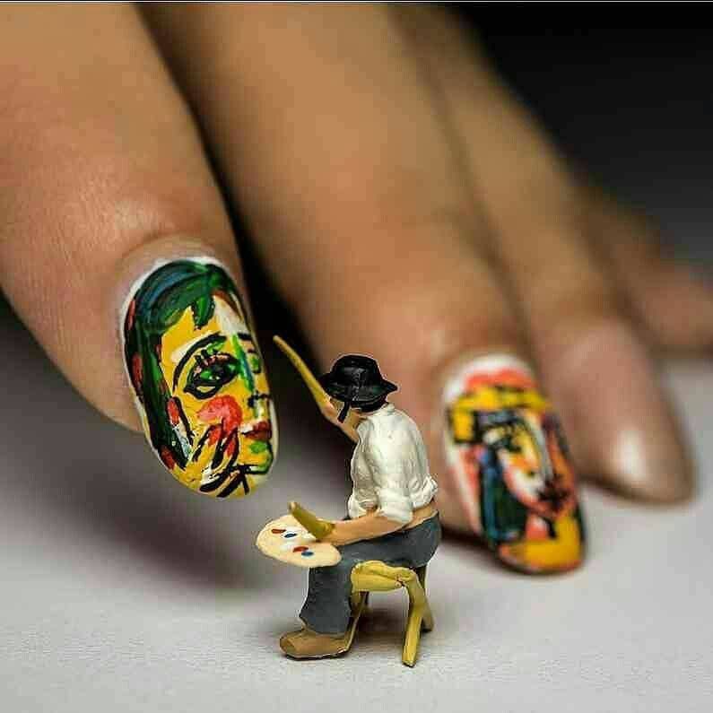 Крупный план ногтей с яркими портретами; рядом миниатюрный художник на стуле с палитрой рисует на ногтях.