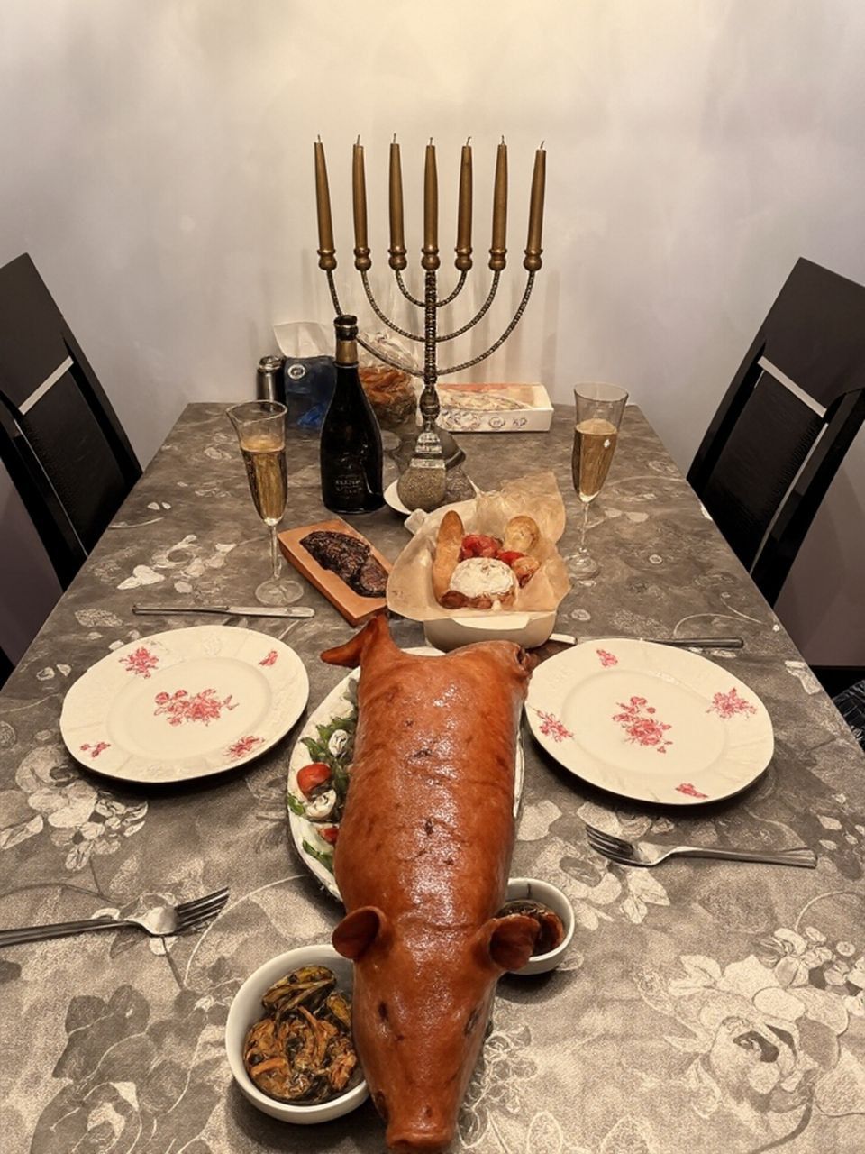 Праздничный стол: запеченная свиная голова на блюде, бокалы с вином, хлеб и десерты, в центре менора.