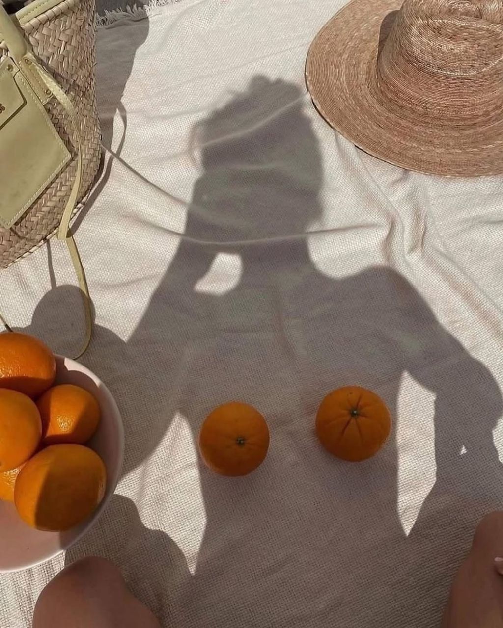 Солнечный пикник: тень человека на тканевом покрывале, апельсины и шляпа рядом.