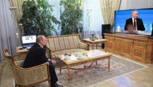 Человек сидит в гостиной перед телевизором, на экране выступает человек в костюме. Между ними ноутбук, чайный сервиз и мебель в стильной гостиной.