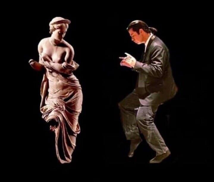 Человек в костюме танцует рядом со статуей на темном фоне.