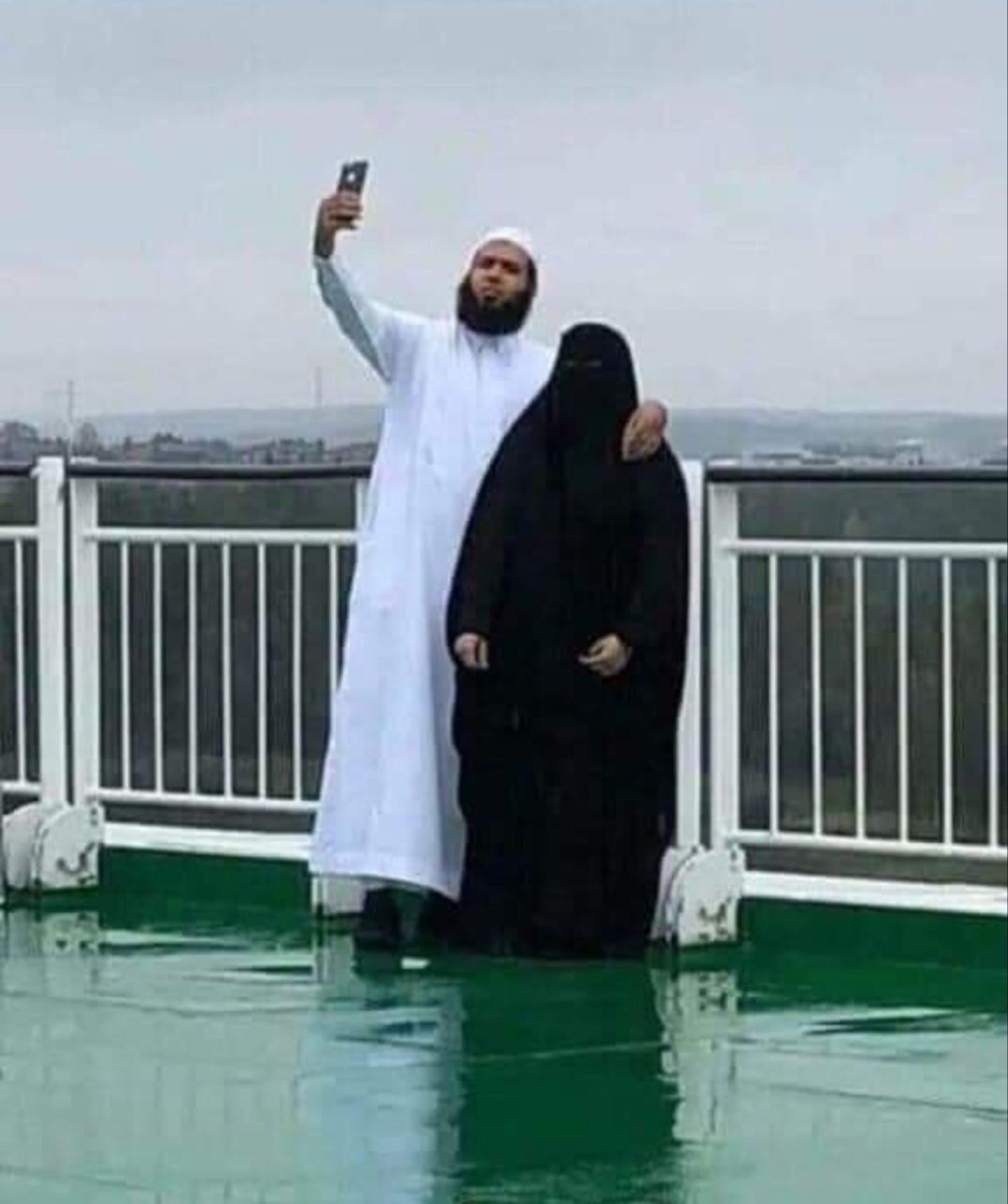 Мужчина в белой робе делает селфи рядом с женщиной в черном чадре на крыше здания.