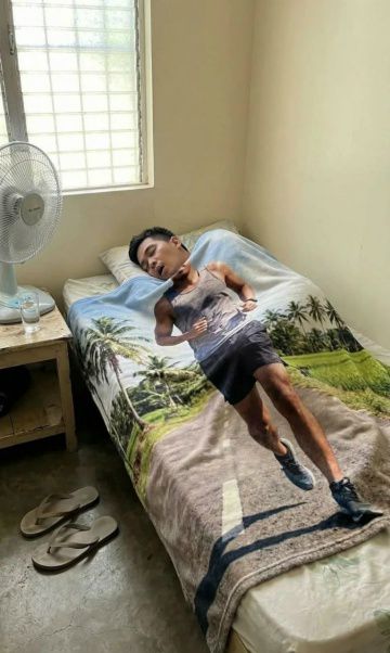 Мужчина спит в кровати под одеялом с рисунком бегущего спортсмена; рядом вентилятор и тапочки.