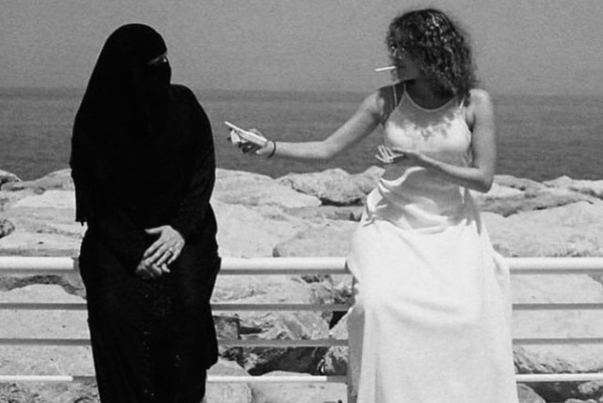 Две женщины у моря: одна в черной абайе, другая в белом платье курит.