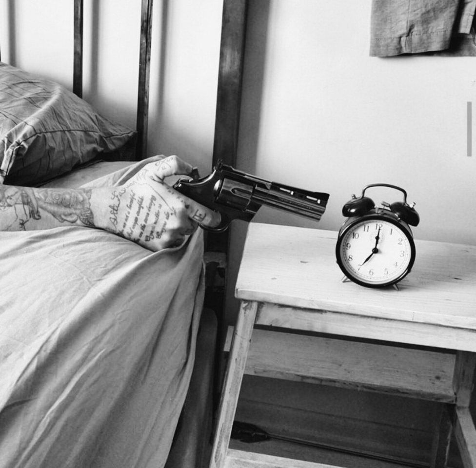 Черно-белая сцена: человек в постели держит пистолет, целясь в будильник на тумбочке.