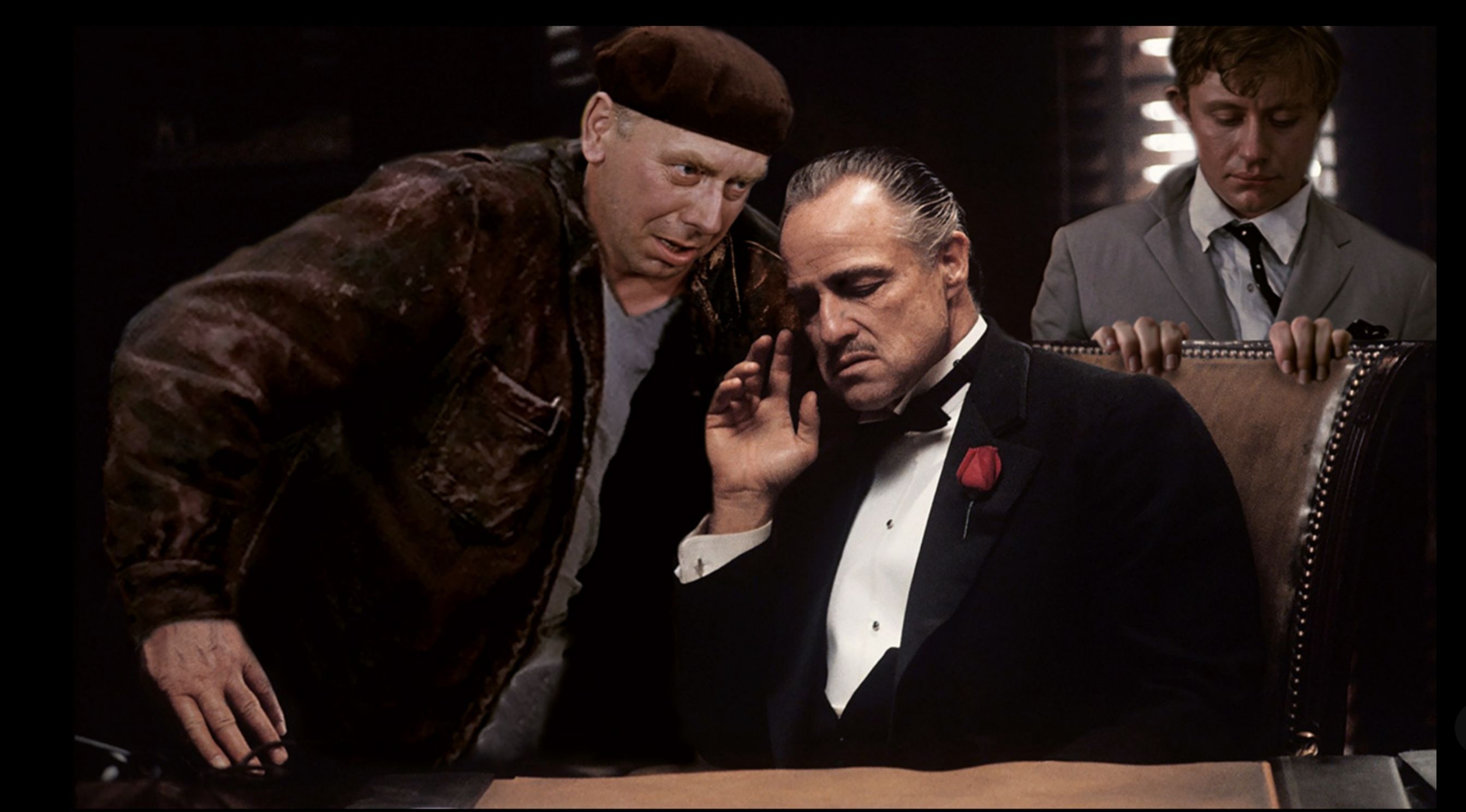 Три мужчины в сцене за столом; один в смокинге держит телефон, рядом стоят двое мужчин, один в кепке и пальто.