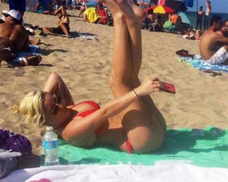 На песчаном пляже женщина в красном купальнике лежит на зелёном коврике и поднимает ноги вверх; вокруг много отдыхающих.