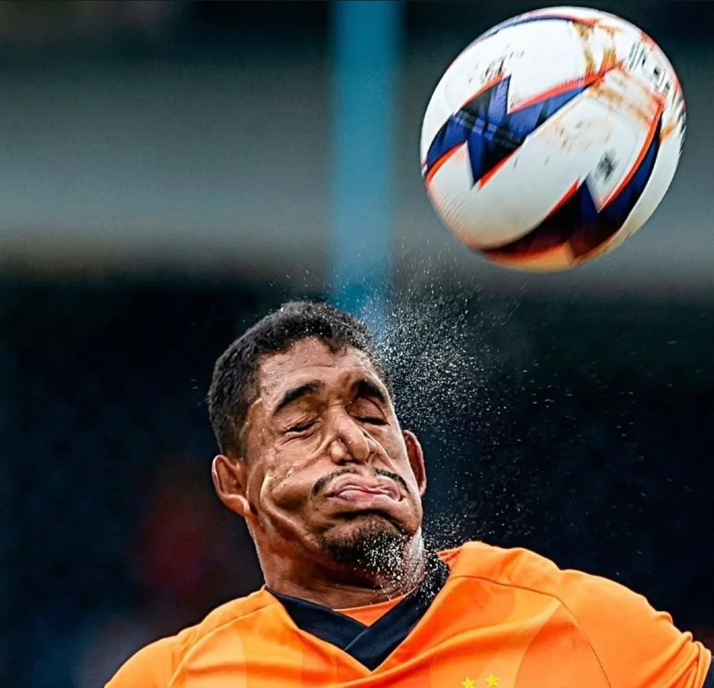 Игрок в оранжевой форме ловит удар мячом, комичный лицевой момент.