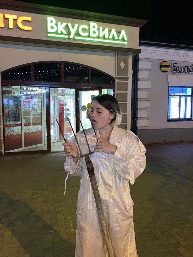 Женщина в светлом платье возле магазина ВкусВилл держит длинный предмет с двумя вилкоподобными концами.