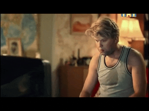 Молодой мужчина в белой майке без рукавов сидит у телевизора в уютной комнате, рядом лампа и карта на стене.