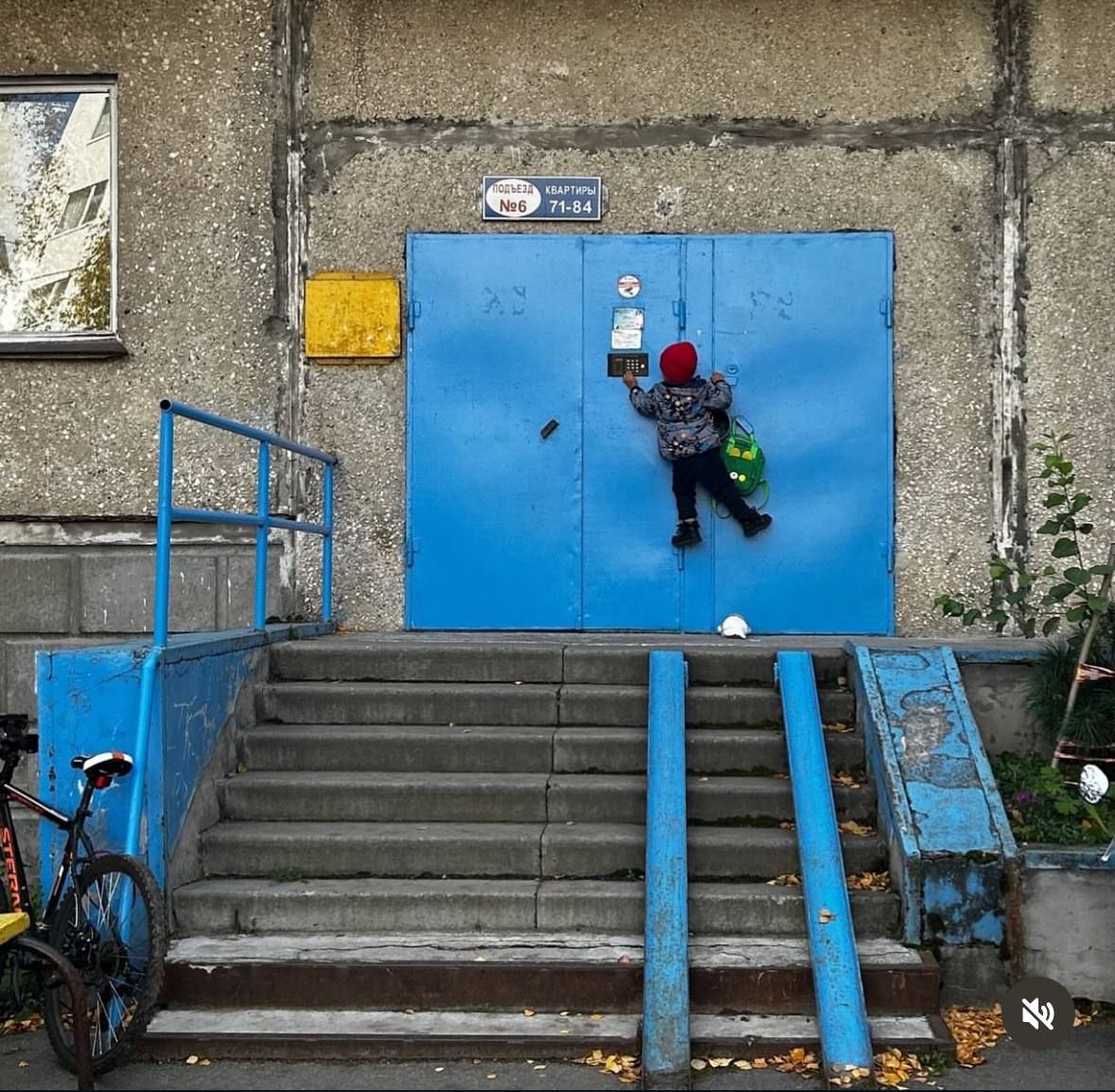 Мальчик в красной шапке цепляется за голубые двери у подъезда возле ступенек и перил.