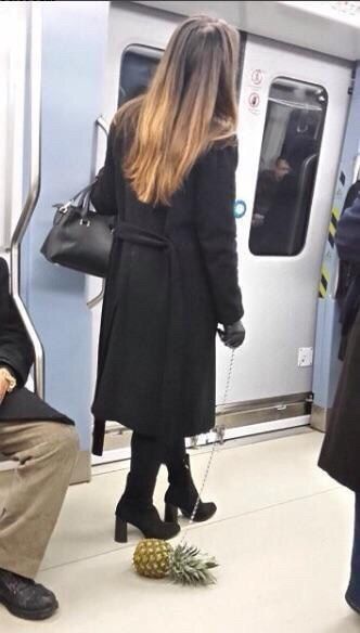 Женщина в черном плаще в метро, рядом на полу лежит ананас на цепочке, как будто предмет для тики.