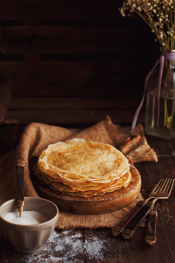 Стопка тонких блинов на деревянной доске в уютной светлой композиции с сахарной пудрой и венчиком на столе.