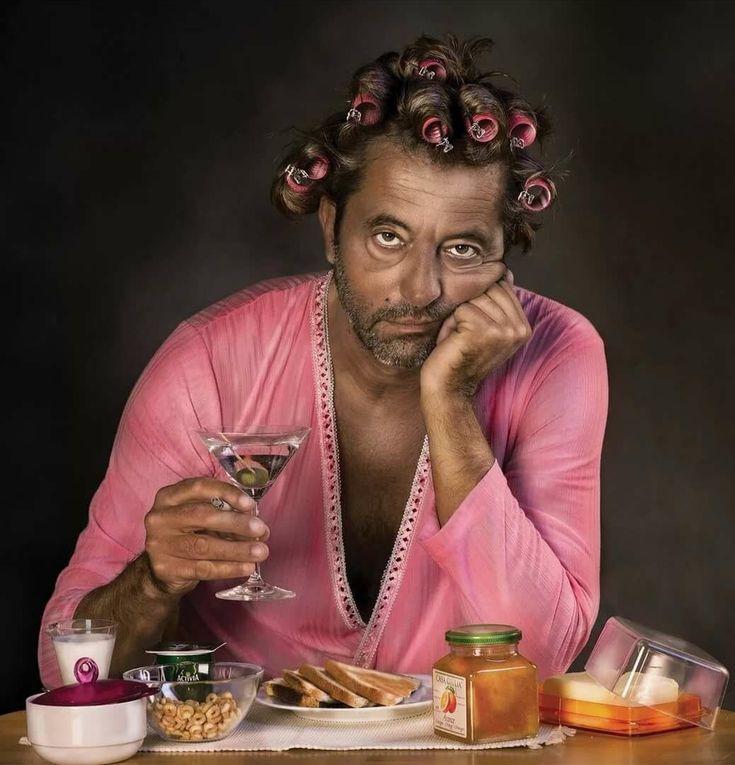Мужчина в розовом халате с бигуди и бокалом на столе с едой