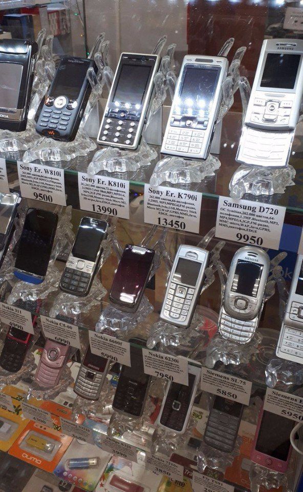 На витрине магазина представлено множество старых мобильников: Sony Ericsson K810i/K790i, W580i, Samsung D720 и другие модели. Видны ценники: 13950, 13450, 9950 и т.д.