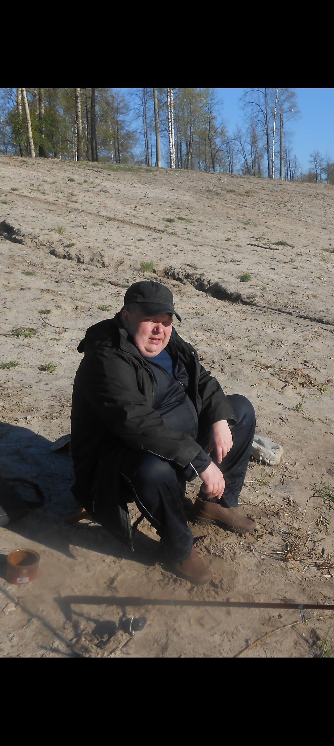 Мужчина в кепке сидит на песчаном берегу, рядом удочка и чашка.