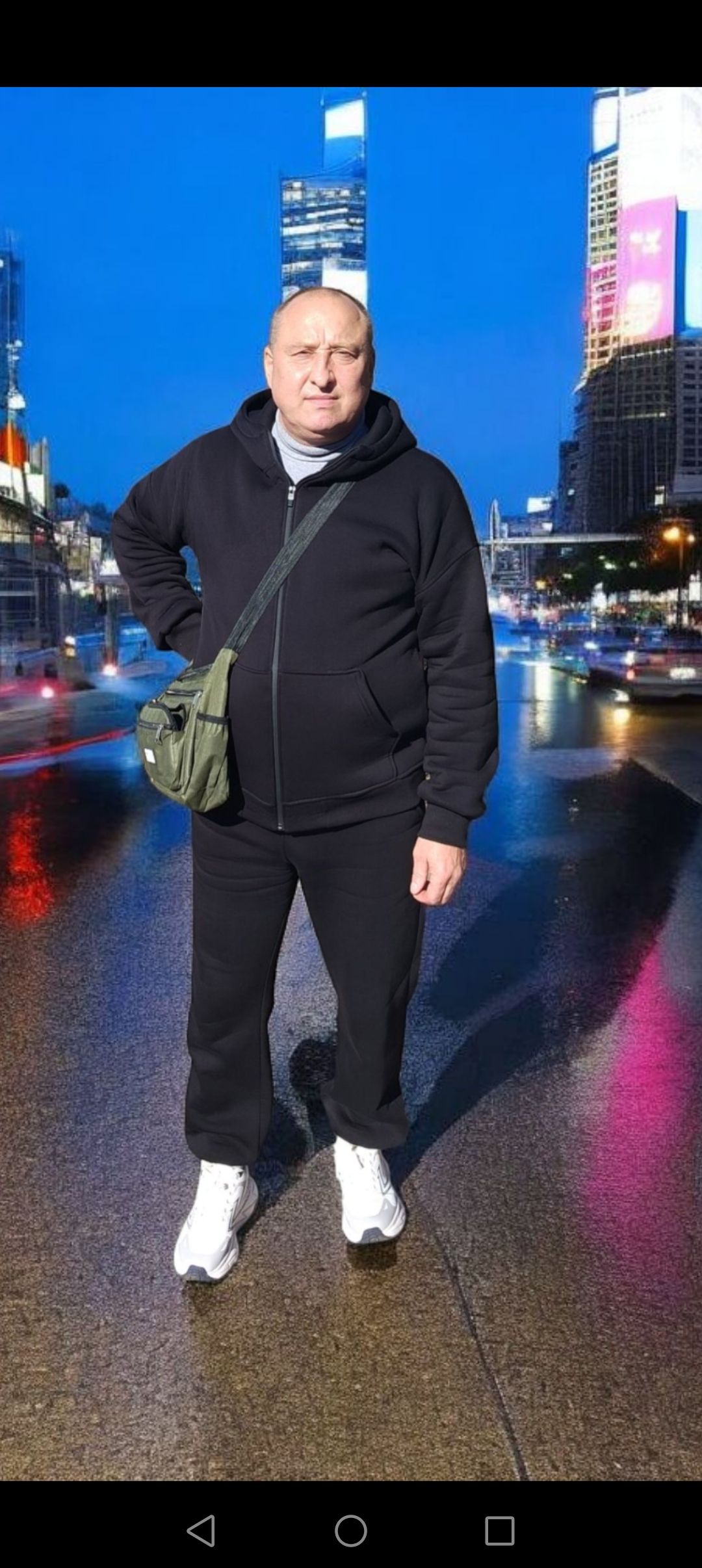 Мужчина в черном костюме и белых кроссовках стоит на мокром городском проспекте с неоновыми огнями на заднем плане.