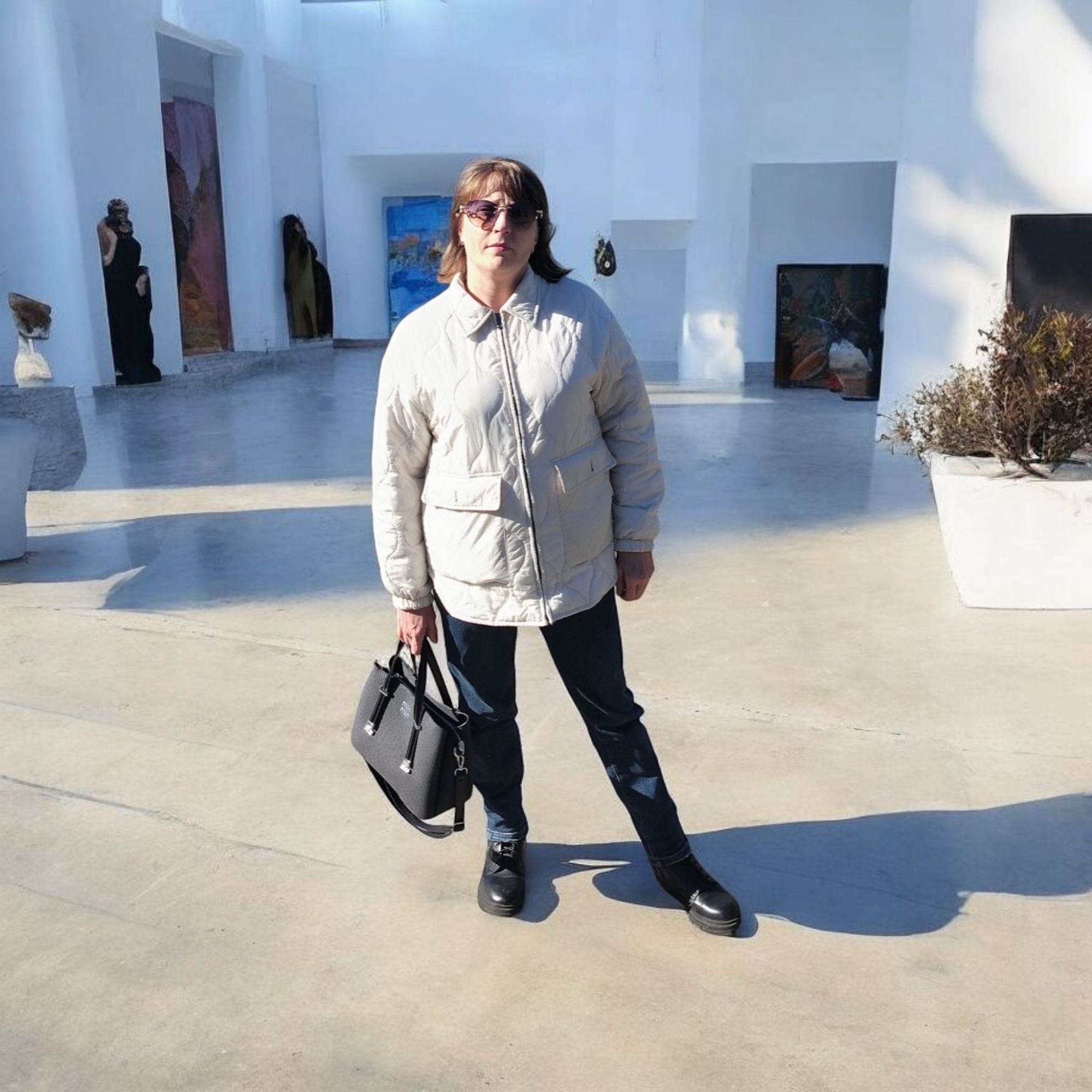 Женщина в светлой куртке на открытой площадке с современным интерьером и скульптурами.