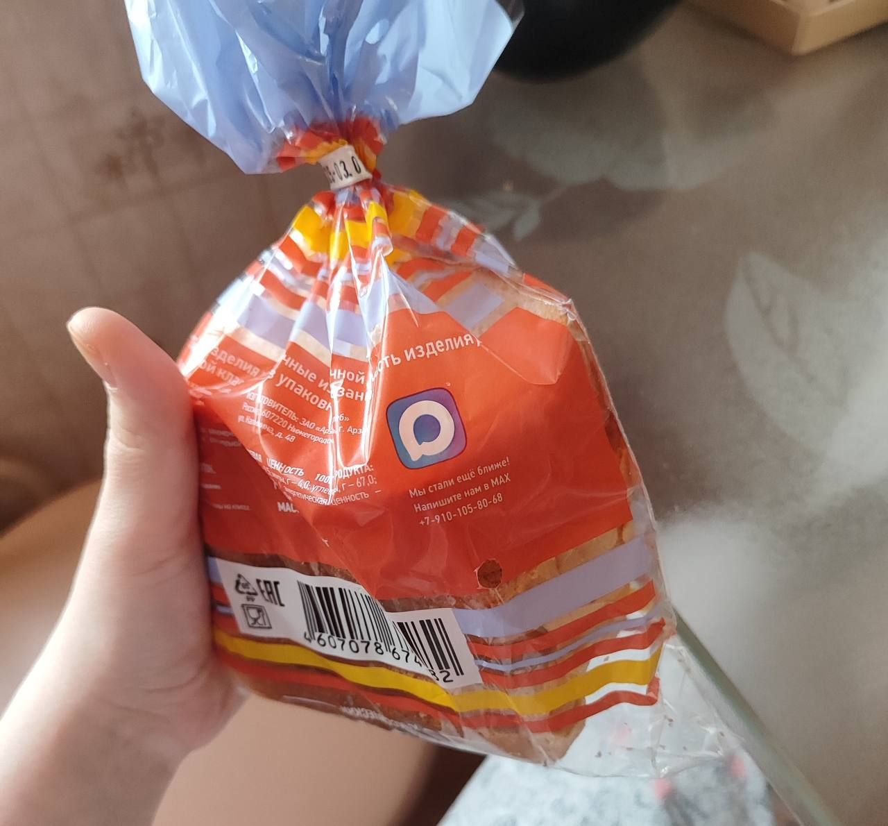 Руку держит ярко-оранжевая запакованная булочка/хлеб в прозрачной обертке.