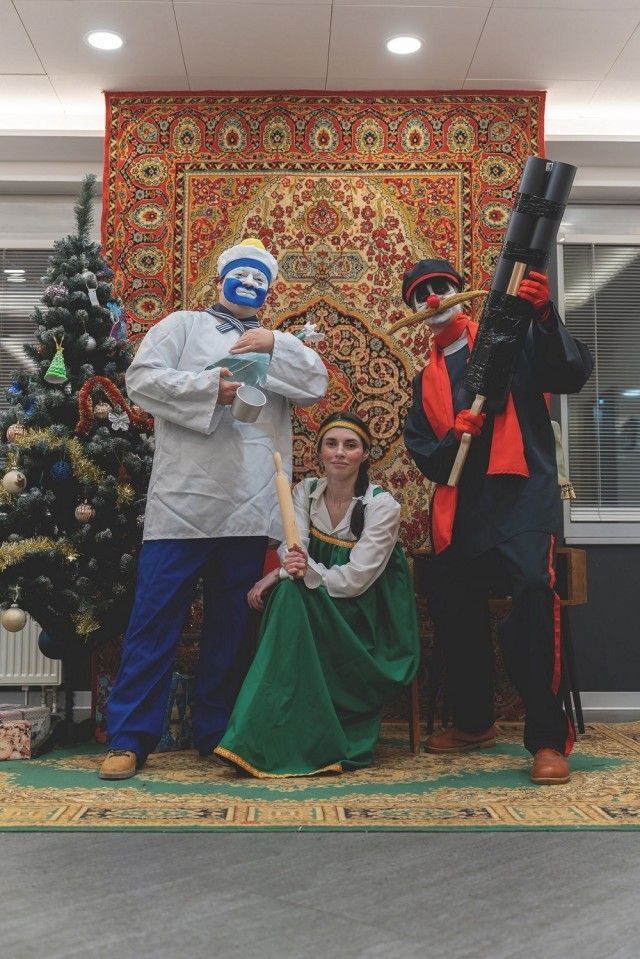 Группа людей в праздничных костюмах на ковре рядом с елкой и ярким тканевым фоном