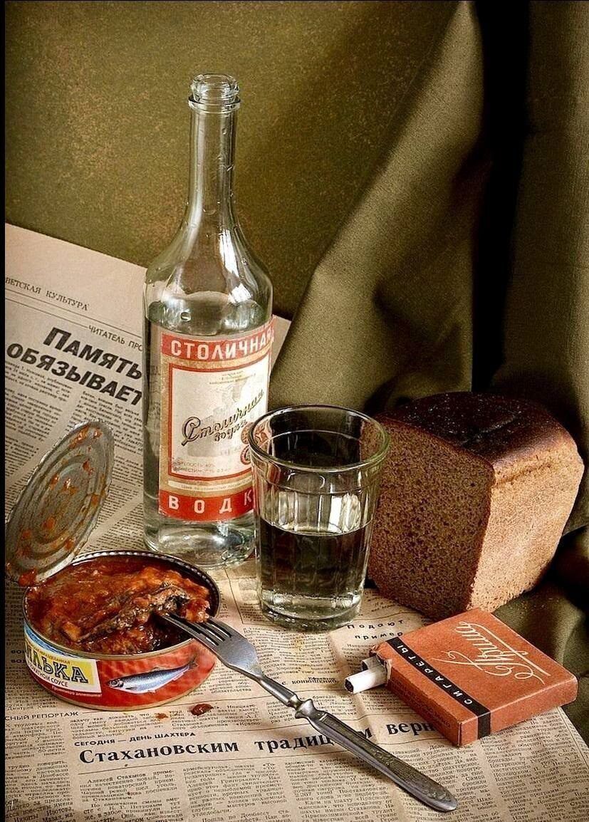 Столичная водка, стакан воды, хлеб, консервы и шоколадка лежат на газетах. На бутылке надпись Столиная, рядом ломтик хлеба и открытая консерва. Всё создает ностальгическую сцену.