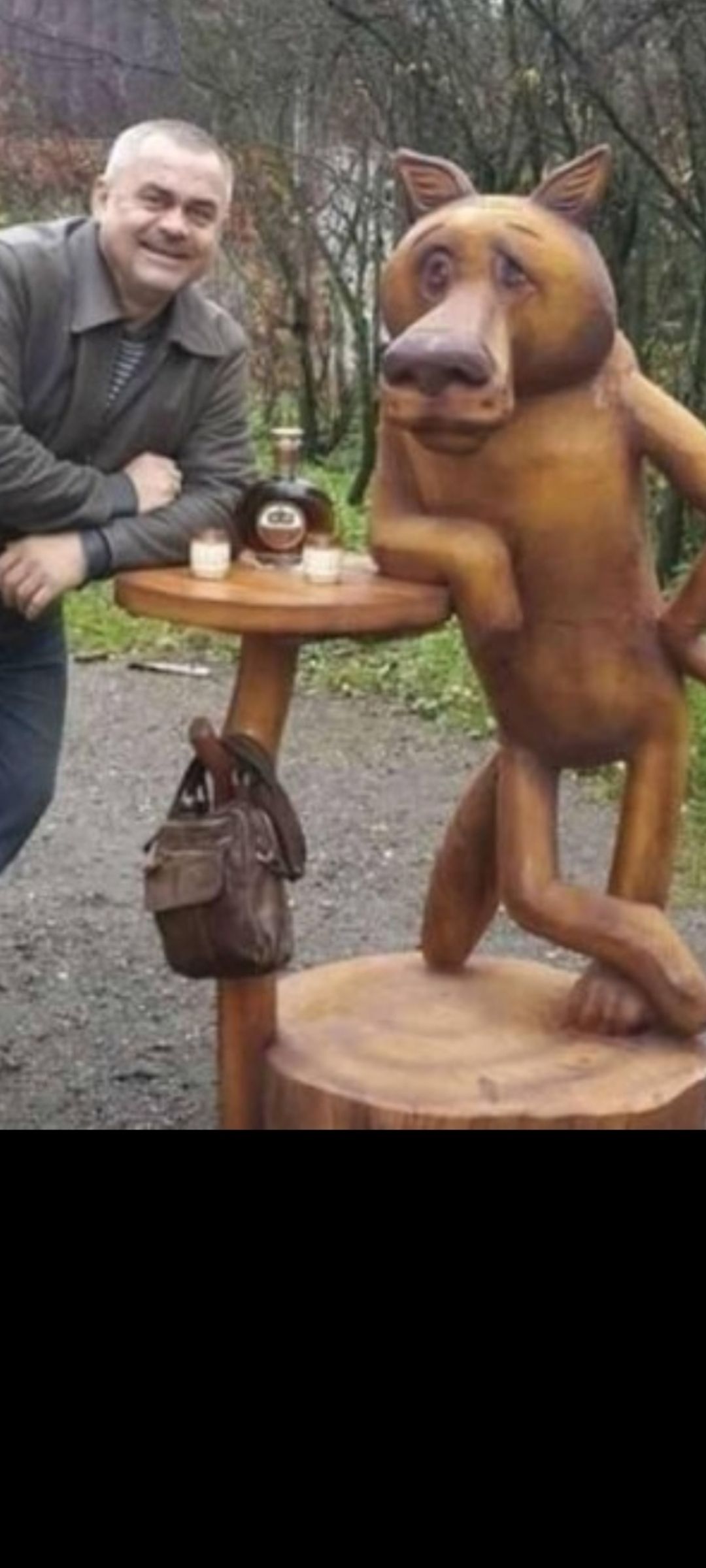Мужчина рядом с резной фигурой свиньи у деревянного столика.