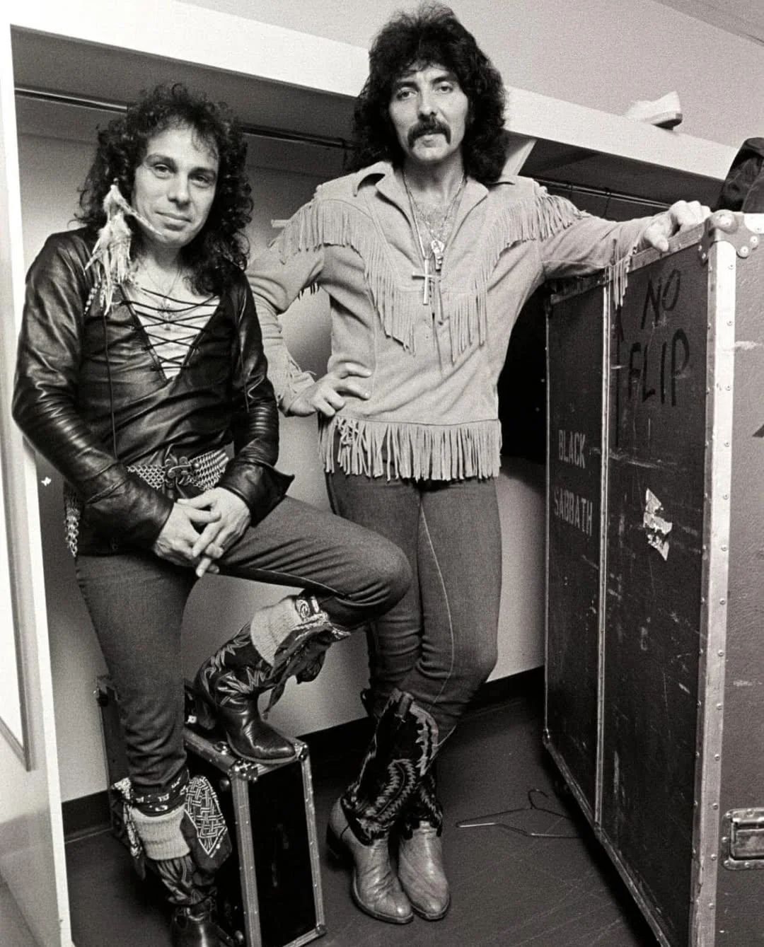 Два рок-музыканта в 70-х стилях позируют в гримёрке рядом с дорожной стойкой.
