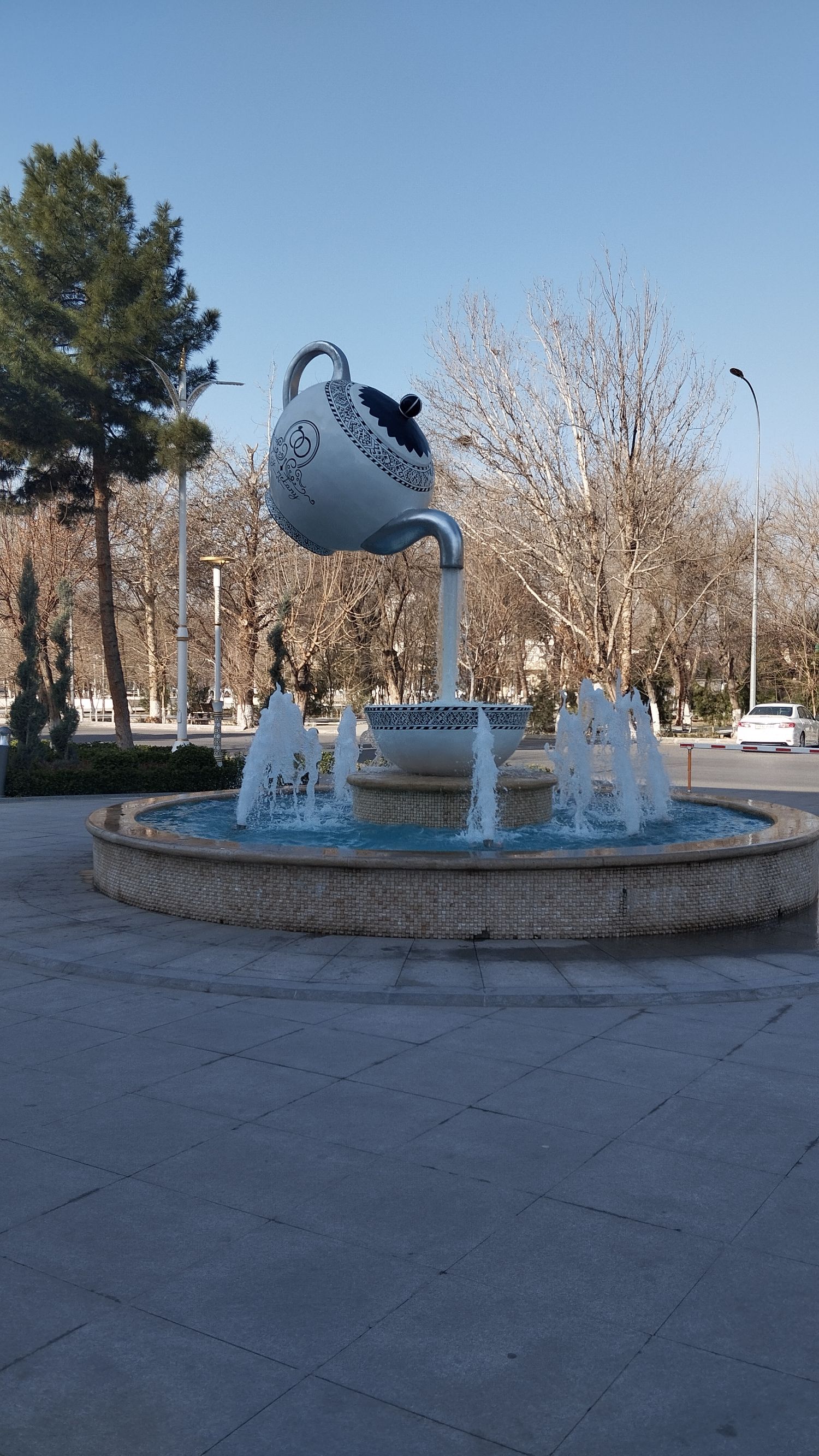 Фонтан в парке: большая чаша воды с гигантским подвесным чайником над ней.