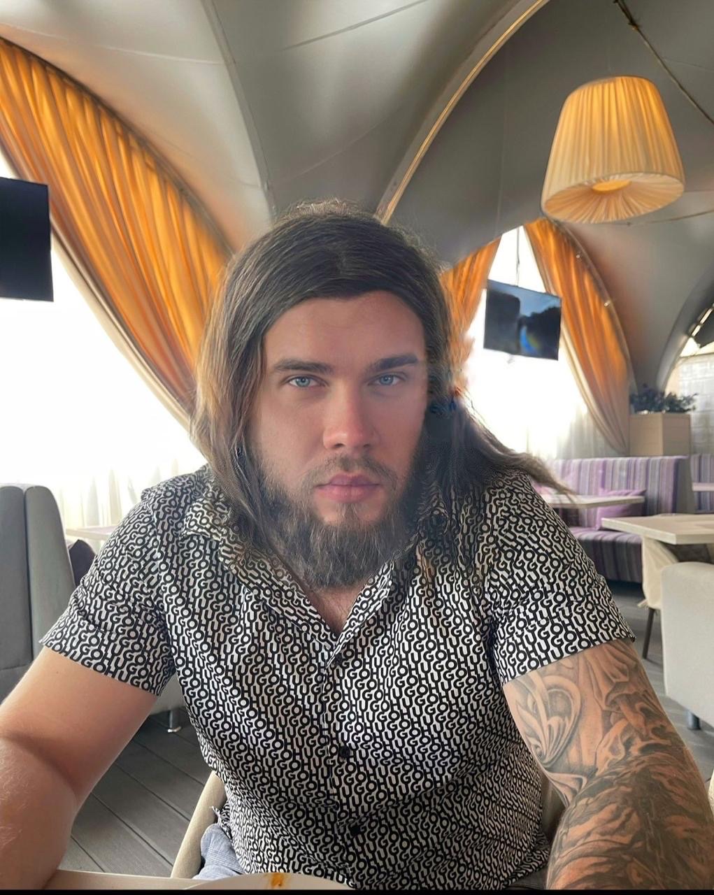 Фотография мужчины с длинными волосами и бородой в светлом помещении с желтыми занавесками.