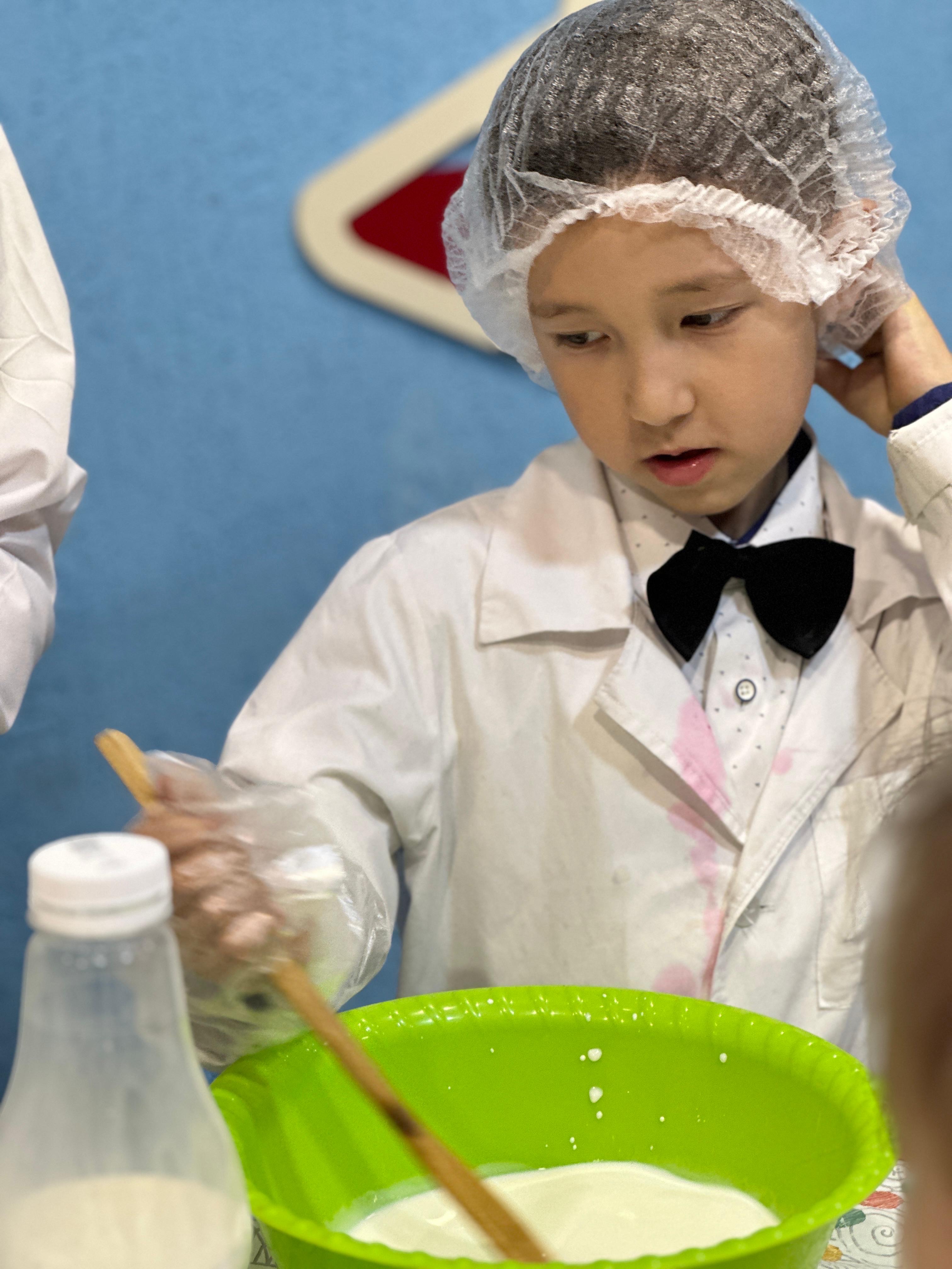 Мальчик в халате и сетке на голове перемешивает ингредиенты в зелёной миске.